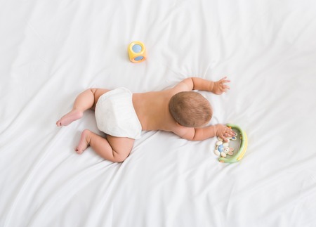 Small baby in diaper lying on his belly on white bed with toys, top view, copy spaceの写真素材