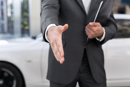 Male car salesman stretching hand for handshake with automobile on backgroundの写真素材