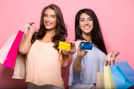 Two happy young women holding credit cards, posing with shopping bags and looking at camera, pink background.の写真素材