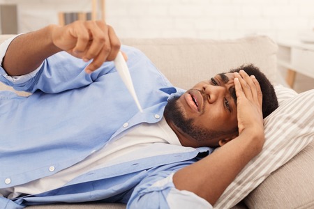 Sick african-american man lying on sofa and checking his temperature with thermometerの写真素材