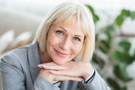 Lovely middle-aged blond woman with beaming smile sitting on sofa and looking at cameraの写真素材