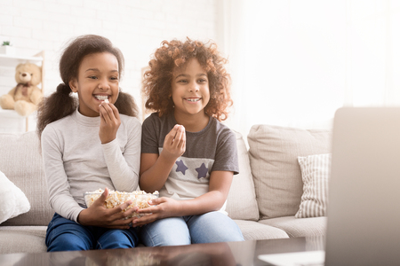 Little girls watching cartoon and eating popcorn, sitting on sofa at homeの写真素材