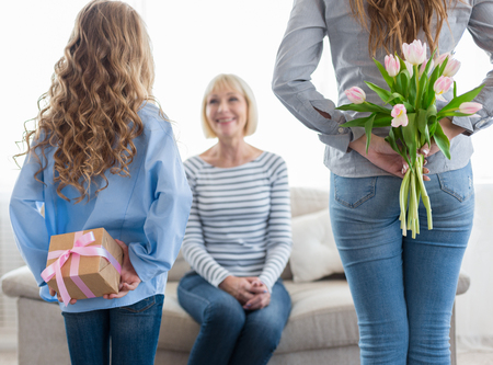 Girl and her mom hiding flowers and gift box for their granny behind backs, wishing happy birthday...