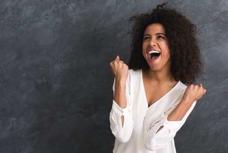 Dark skinned triumphing girl raises fists happily on dark background. We are the champion conceptの写真素材