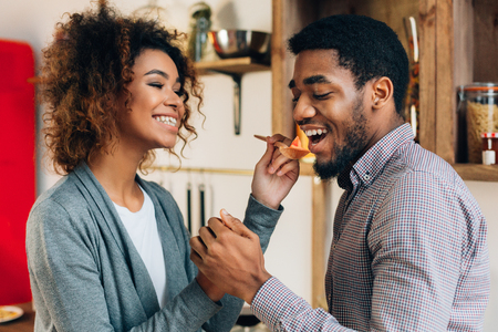 Cooking lunch together. African-american woman in kitchen letting man to taste meal with wooden spoon, closeupの写真素材
