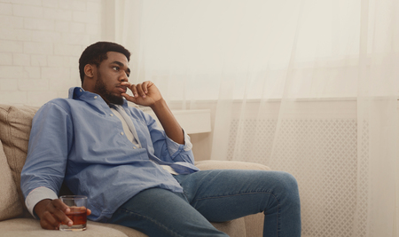 Depression. Young african-american man drinking alcohol at home, feeling lonely and sick, holding glass of whiskey, panorama, copy spaceの写真素材