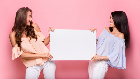 Cheerful women holding blank advertisement sheet and smiling to each other, pink studio background,...