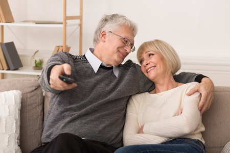 Relaxed senior couple watching television, switching channels by remote control at homeの写真素材