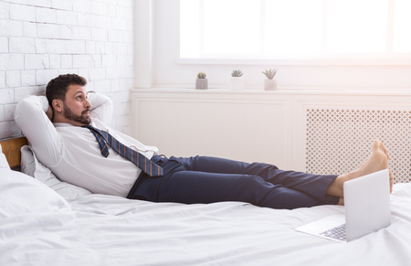 Businessman relaxing from working on laptop, lying on bed in hotel roomの写真素材