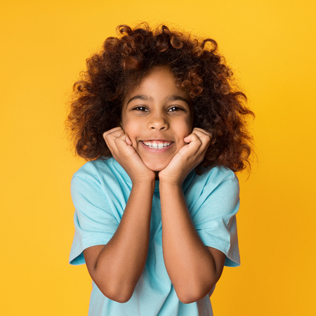Cute african-american girl posing, holding hands under chin over yellow background, crop