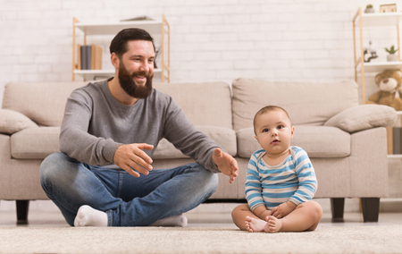 Cheerful father playing with his baby son, sitting on floor at home, copy spaceの写真素材