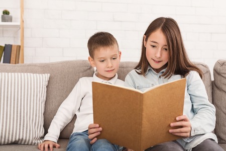 Brother and sister reading book together, sitting on sofaの写真素材