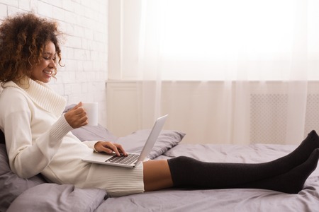African-american girl surfing net on laptop and drinking coffee, sitting on bed, empty spaceの写真素材