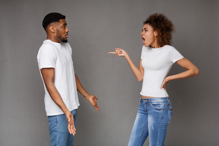 Emotional african-american girl screaming at her confused boyfriend, gray studio background.の写真素材