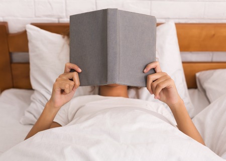 Woman reading book and covering face, lying in bed in the morningの写真素材