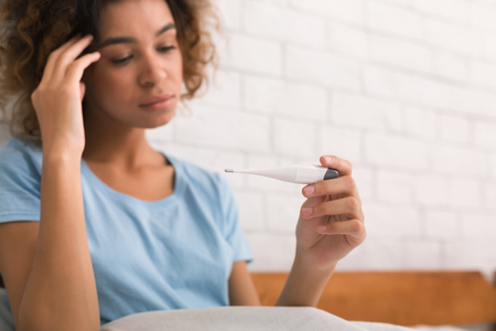 Woman with flu virus measuring her temperature with thermometer, lying in bedの写真素材
