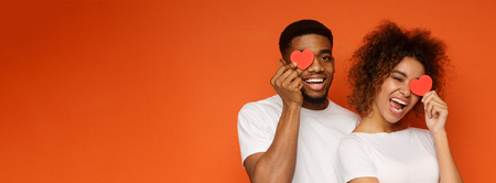 Young cheerful african-american couple in love holding red hearts over eyes and smiling, orange panorama background with empty spaceの写真素材