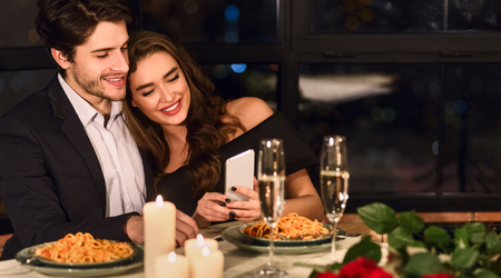 Happy couple looking photos on smartphone during romantic dating in restaurantの写真素材
