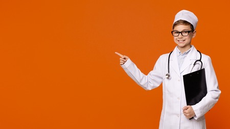 Cute smiling boy playing doctor, pointing away at empty space on orange panorama backgroundの写真素材