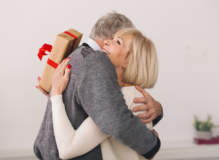 Mature man giving present to wife, hugging and smiling at homeの写真素材