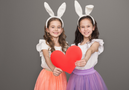Family love and sisterhood. Two happy little girl in bunny ears holding big red paper heart on gray backgroundの写真素材