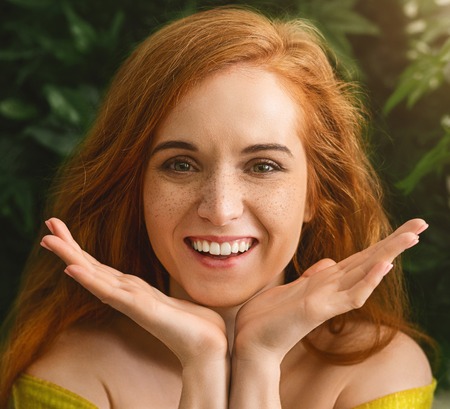 Cheerful foxy girl enjoying her freckles, holding face in palms, smiling at cameraの写真素材