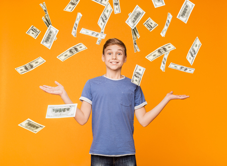 Cheerful successful boy standing under money shower, enjoying falling dollar banknotes on orange backgroundの写真素材