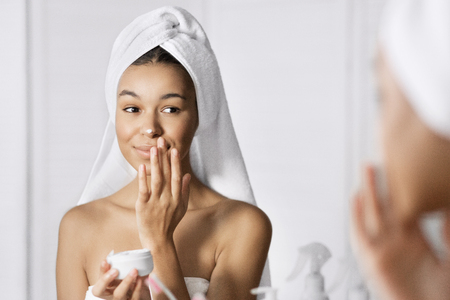 Cute afro-american girl applying cream on her face in bathroom, looking in mirror, reflectingの写真素材