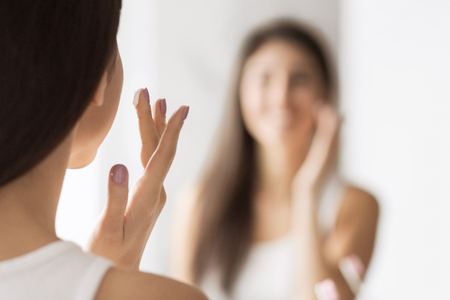 Beautiful young woman applying cream on her face, standing in bathroom, reflecting in mirror. Everyday skincare conceptの写真素材