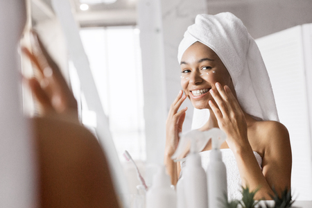 Smiling afro girl after shower with towel on head in bathroom looking in mirror. doing facial treatment.の写真素材