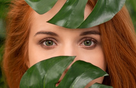 Skin care, pure beauty, cosmetics concept .Young green-eyed redhead girl looking through monstera...