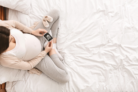 Pregnant woman looking at her baby sonography, enjoying future motherhood with first photo of her unborn child and tiny shoes, top view with free spaceの写真素材