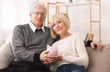 Family budget. Senior couple holding piggybank and looking at camera at homeの写真素材