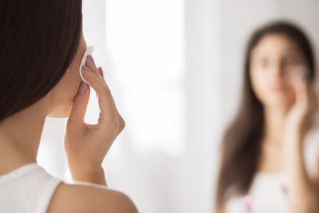 Young woman cleaning face with cotton pad in front of mirror in bathroom. Healthy pure skin conceptの写真素材
