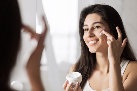 Happy girl applying cream on her face, standing in bathroom, reflecting in mirror. Vitamin care for young skin conceptの写真素材