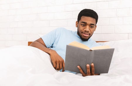 Comfortable weekend. Young african-american guy enjoying book, lying in bed, free spaceの写真素材