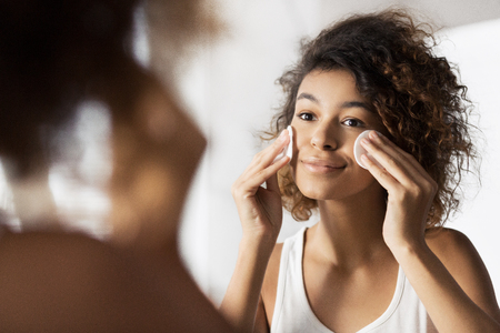 Beautiful afro-american girl cleaning face with cotton pads looking in mirror at bathroom. Daily makeup removing conceptの写真素材