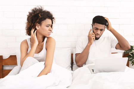 Married couple problem and relationship communication issue. Young attractive woman feeling unsatisfied and frustrated in bed with husband, man workong on laptop and ignoring herの写真素材