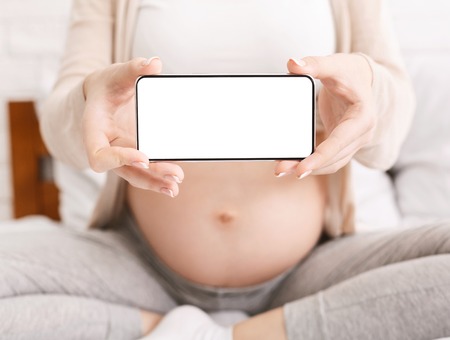 Pregnancy calendar application. Pregnant woman holding cellphone with blank screen near her big...