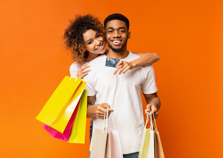Cheerful african-american couple of shopaholics satisfied with their bank, embracing with credit card and shopping bags on orange backgroundの写真素材