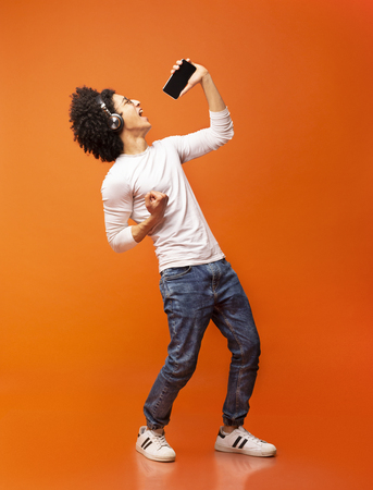 Young emotional black man singing his favorite song, using smartphone as microphone, orange studio backgroundの写真素材