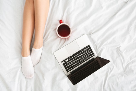 Cozy time in bed. Top view on female legs, laptop and cup of coffee on white sheetsの写真素材