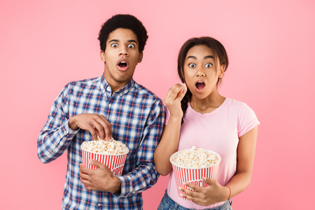 Shocked couple watching scary movie, holding bucket of popcorn on pink backgroundの写真素材