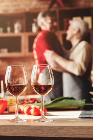 Senior couple in love dancing on their anniversary, celebrating with wine in kitchen, free spaceの写真素材