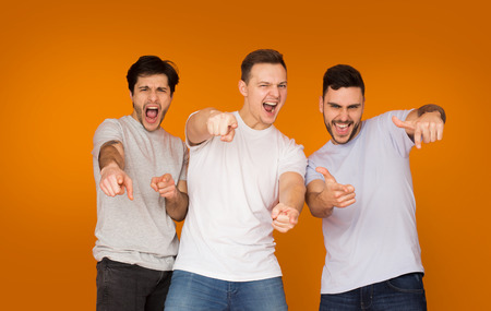 Excited friends pointing fingers at camera over orange background