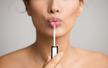 Young woman applying lip gloss on her lips over grey backgroundの写真素材