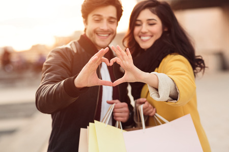 Happy couple showing heart sign with hands, holding shopping bags outdoorsの写真素材