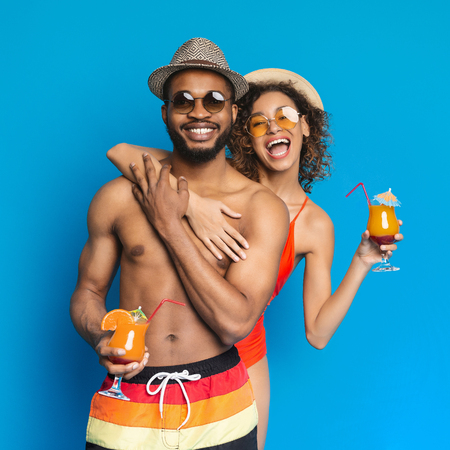 Enjoying summer vacation. Happy african american girl and guy holding cocktails, smiling and embracing on blue studio backgroundの写真素材