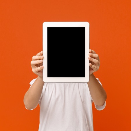 Tablet face. Little boy hiding his face behind portable computer device on orange background, free...