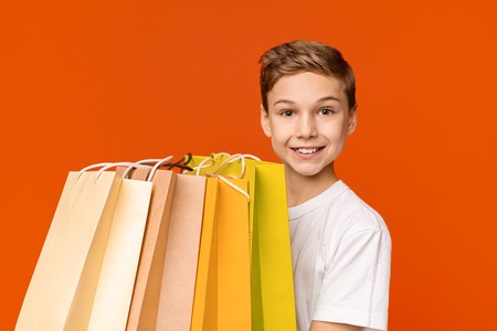 Cheerful teen boy holding lots of shopping bags and smiling, orange studio backgroundの写真素材